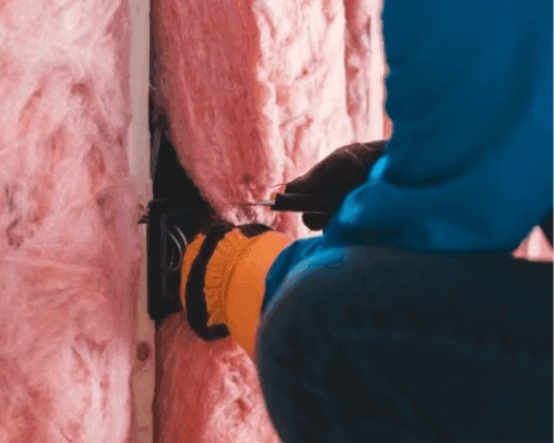 Installer fitting pink fibreglass batts around an electrical box between wall studs